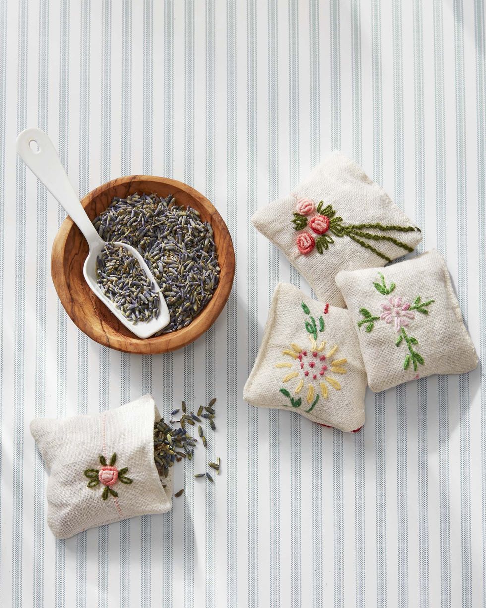 4 small, cotton lavender pouches with a dish and a spoon of lavender are set against a blue and white striped background. The pouches have flowers embroidered on them.