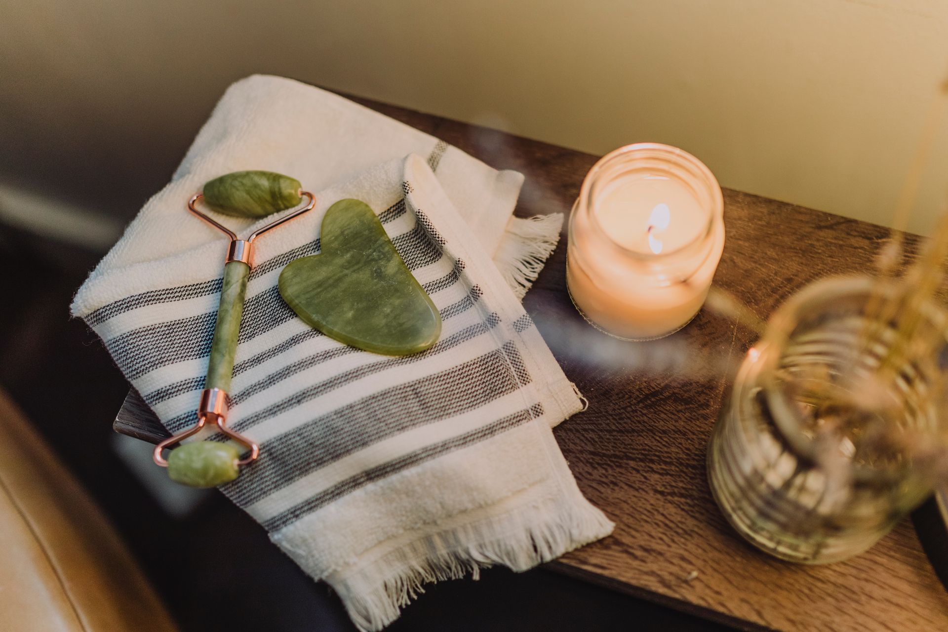 A wooden worktop has a lit candle on top of it, along with a blue and white striped towel, with a green Jade stone and face roller.