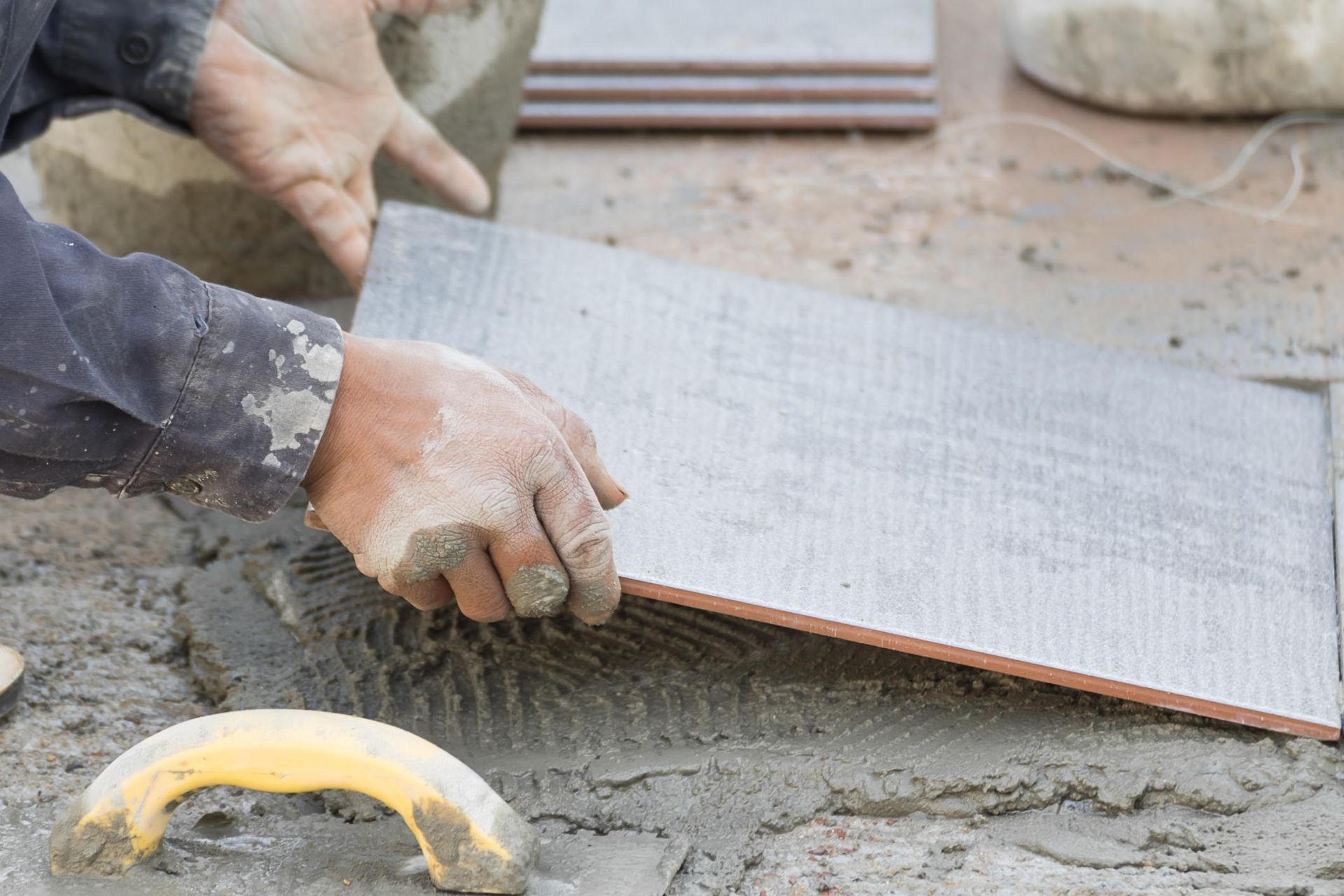 Hands placing a gray tile on wet mortar, with other tiles and a trowel visible.