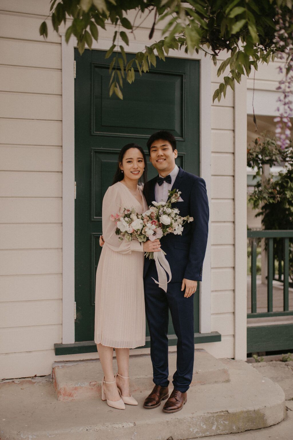 Couple smiles, pose for photo outside white building with green door. Woman holds bouquet, man wears navy suit.