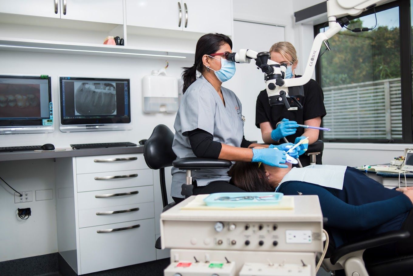 Dentist in pink scrubs pointing at a screen, explaining to a smiling patient in a dental chair.