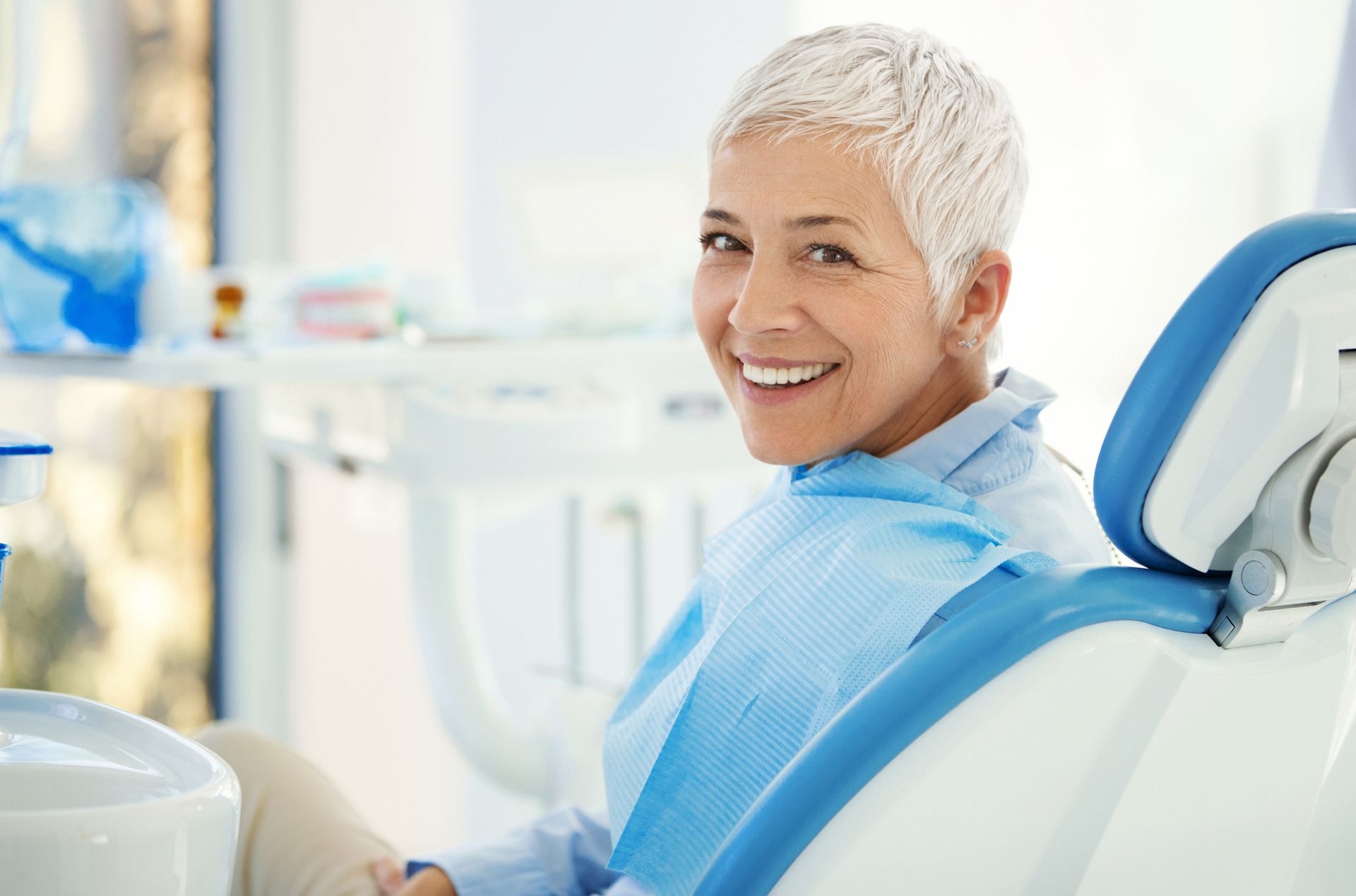 Woman with short, white hair smiles while in a dentist's chair; blue chair and bib.