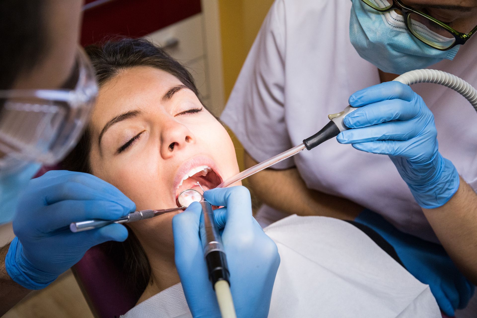 Woman at dentist; two dental professionals working in her mouth with tools.