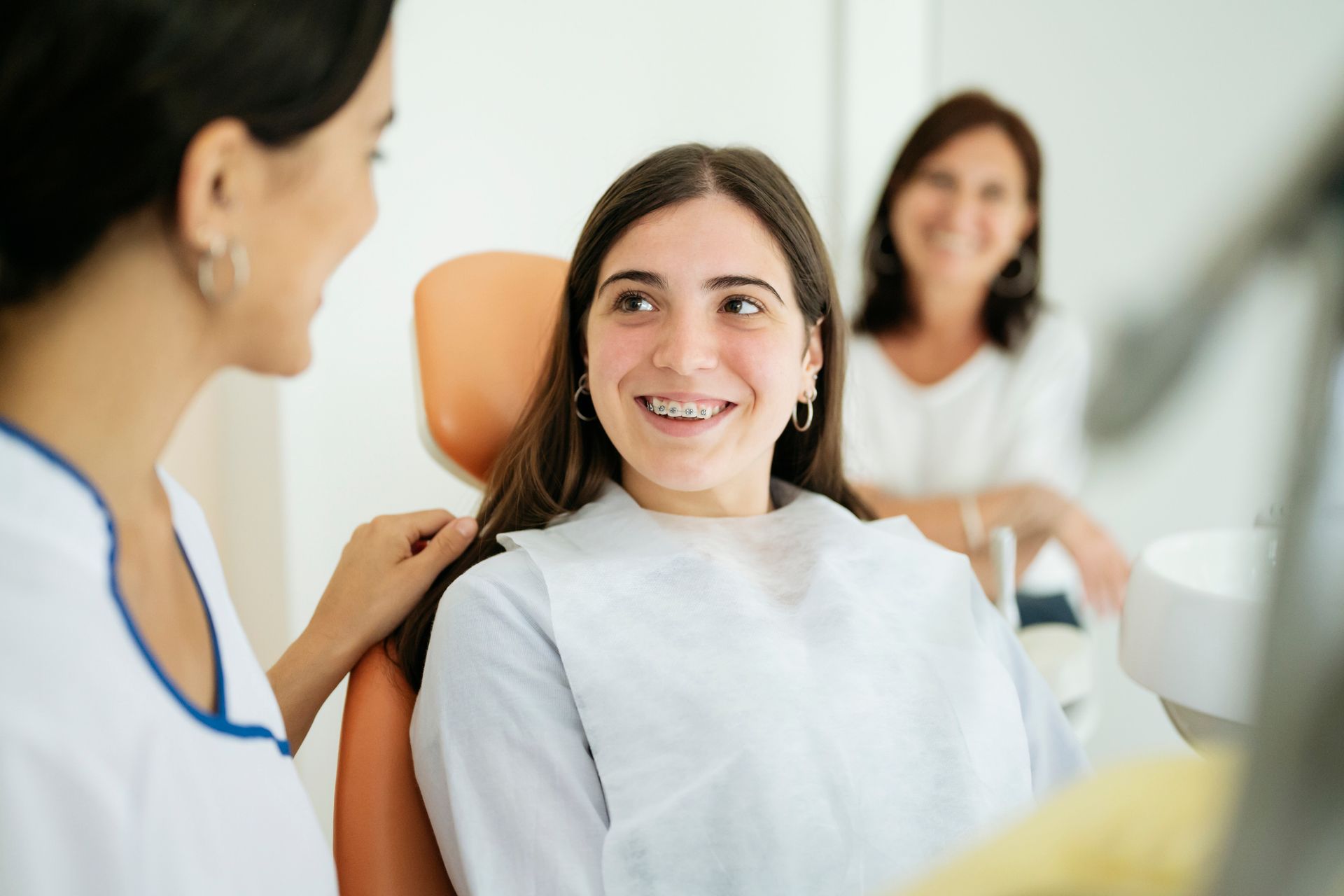 A smiling teen in a dentist's chair, being spoken to by a dental hygienist; a woman smiles in the background.
