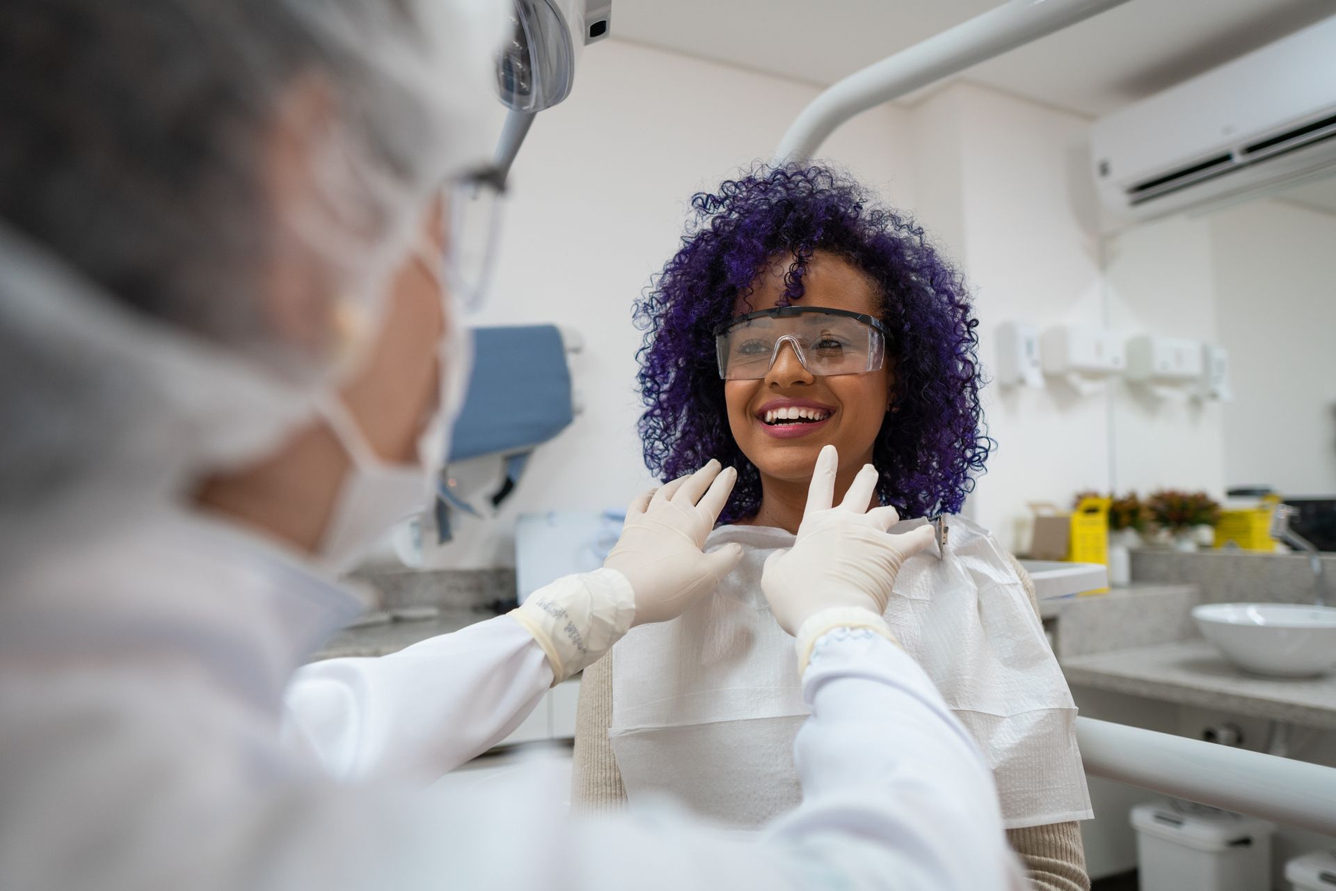 Dentist examining a smiling person with purple hair in a dental office.