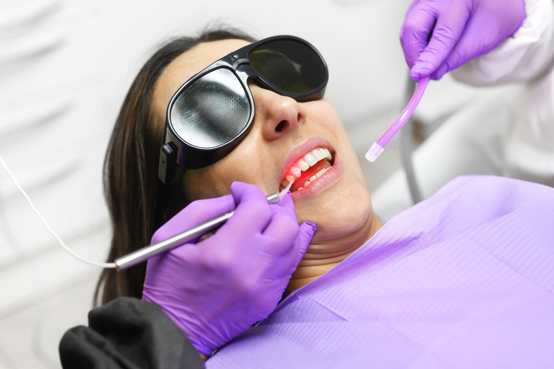 Dentist in pink scrubs pointing at a screen, explaining to a smiling patient in a dental chair.