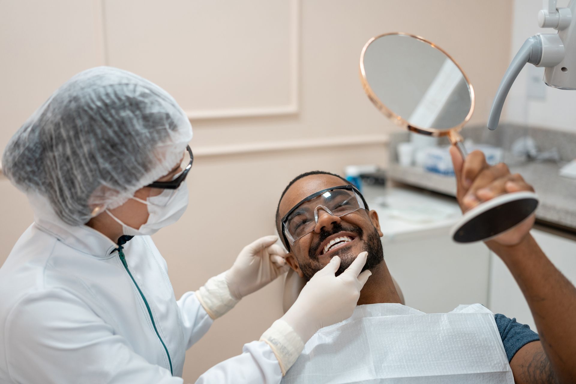 Dentist examining a Black man's teeth, who is looking in a hand mirror. Both are smiling, in a dental office.