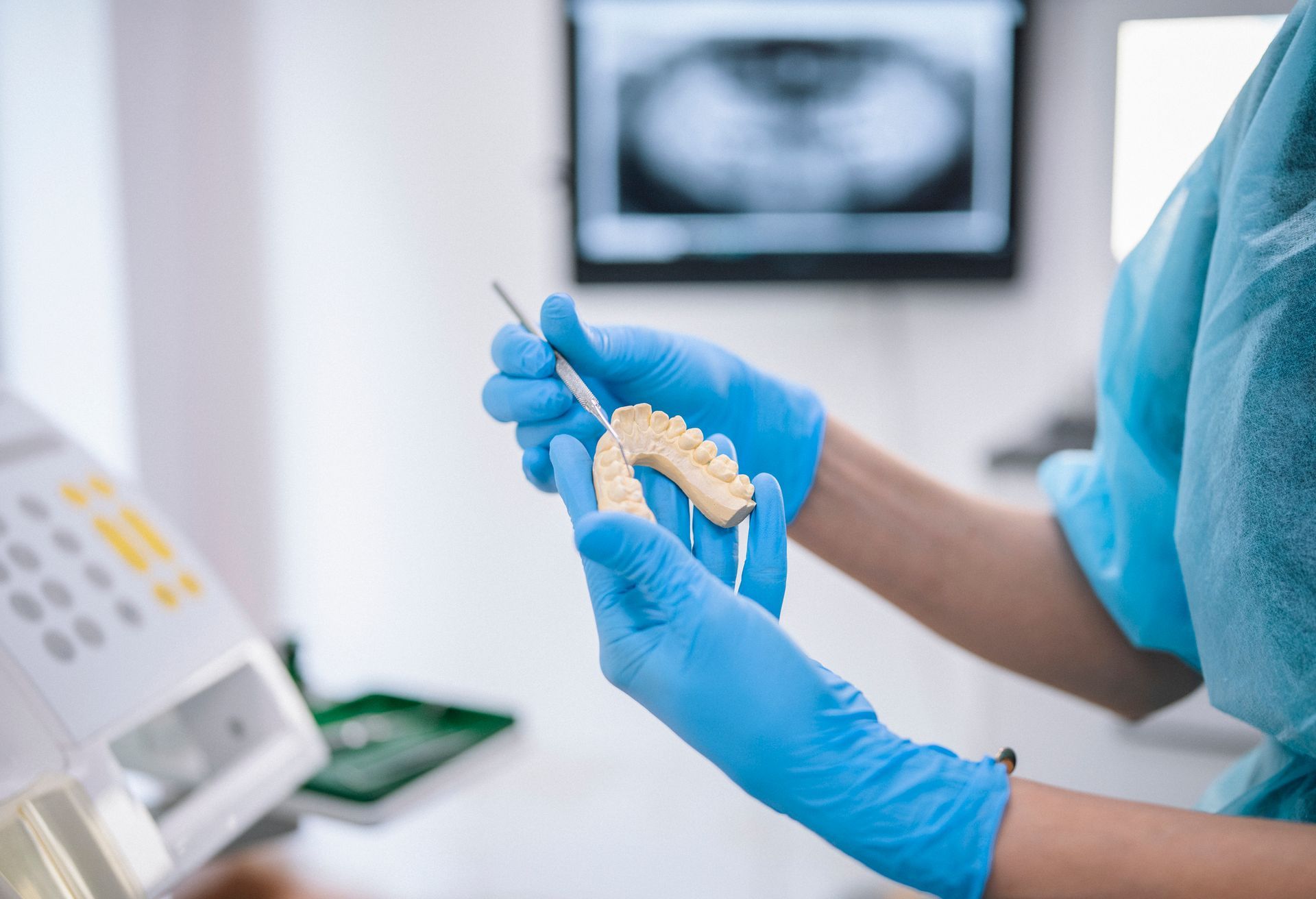 Dentist in blue gloves holding dental model and tool, with X-ray and equipment in the background.