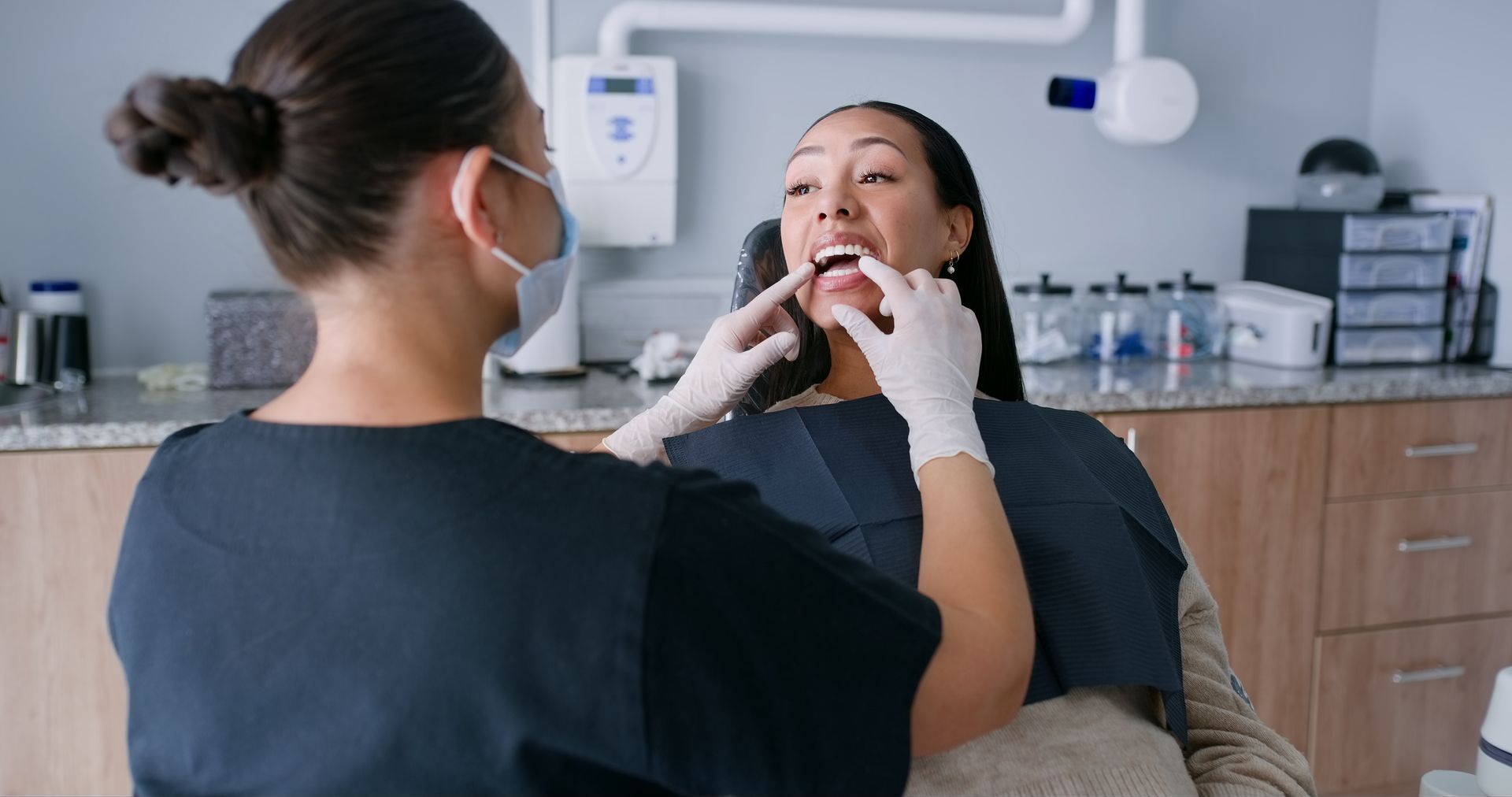 Dentist examines a patient's mouth in a dental office. The dentist wears a mask and gloves. The patient has her mouth open.