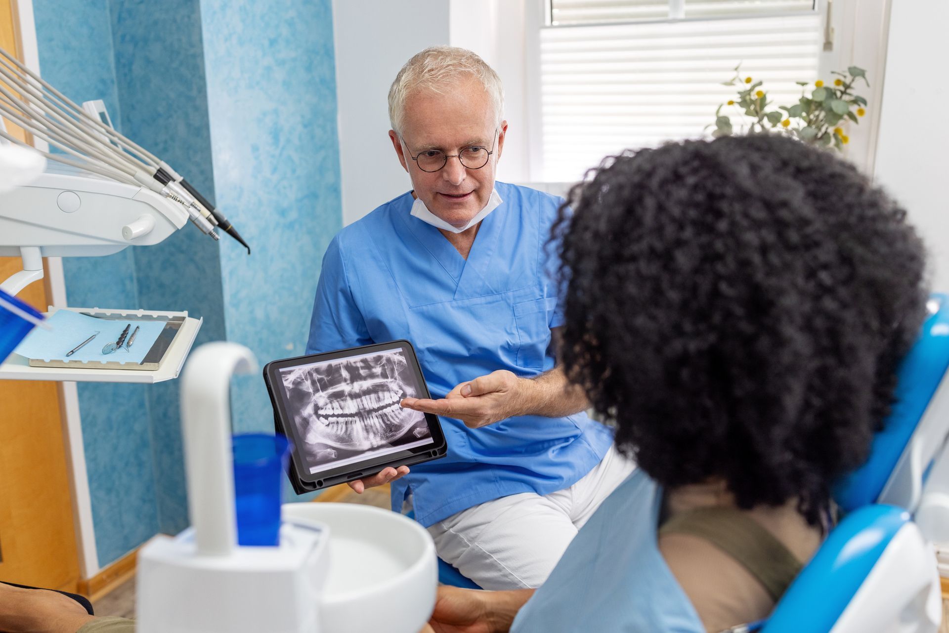 Dentist pointing to dental X-ray on tablet, explaining to a patient. Blue scrubs, dental chair, and light blue wall.