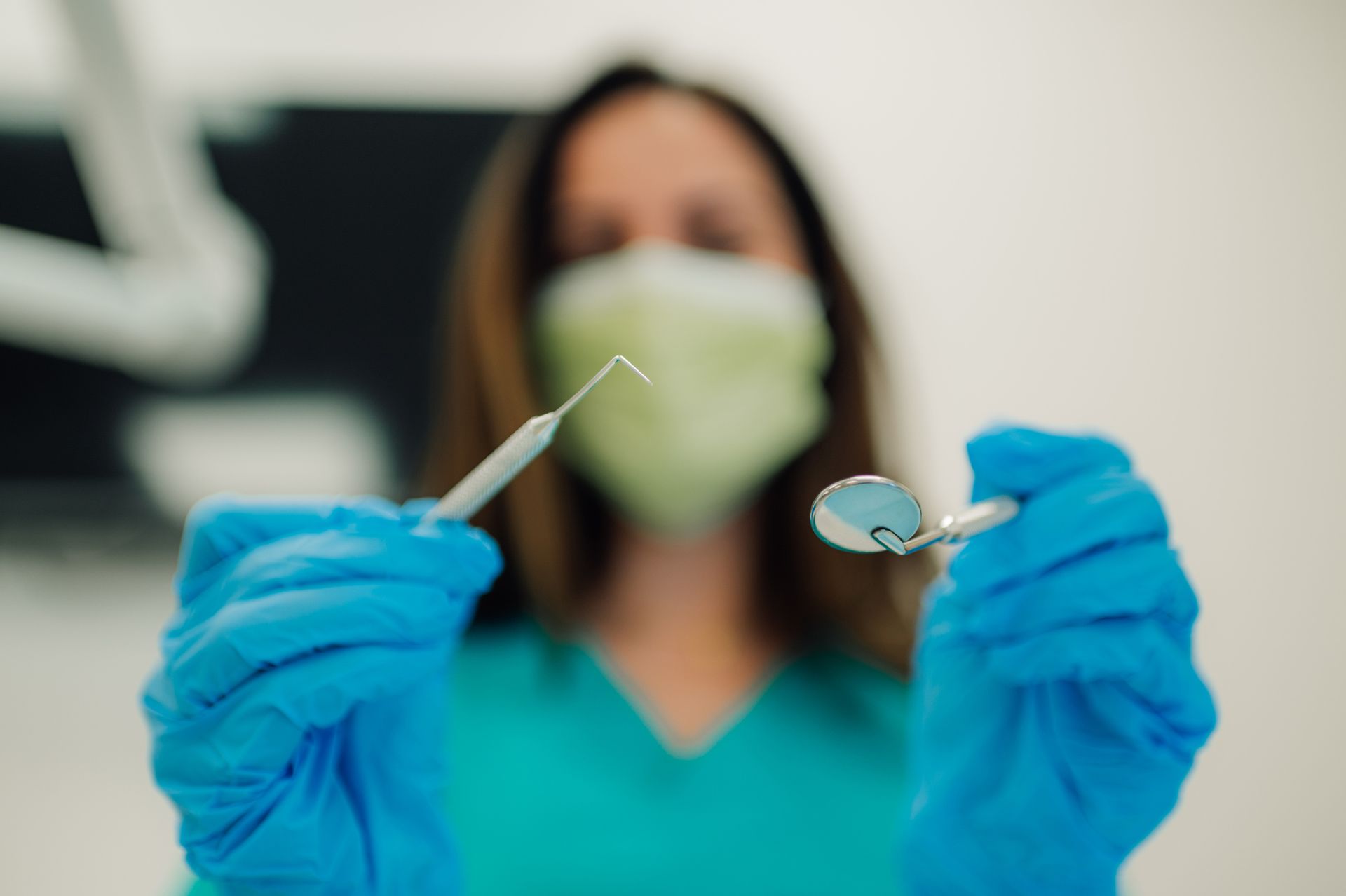 Dentist in blue gloves and scrubs holds dental tools, a probe and mirror, in a medical setting.