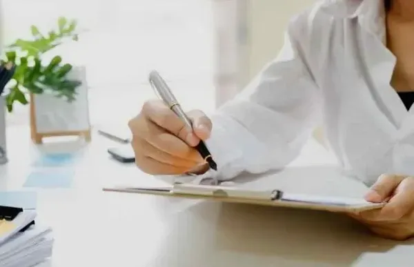 Person in white shirt writing on clipboard with pen at a desk.