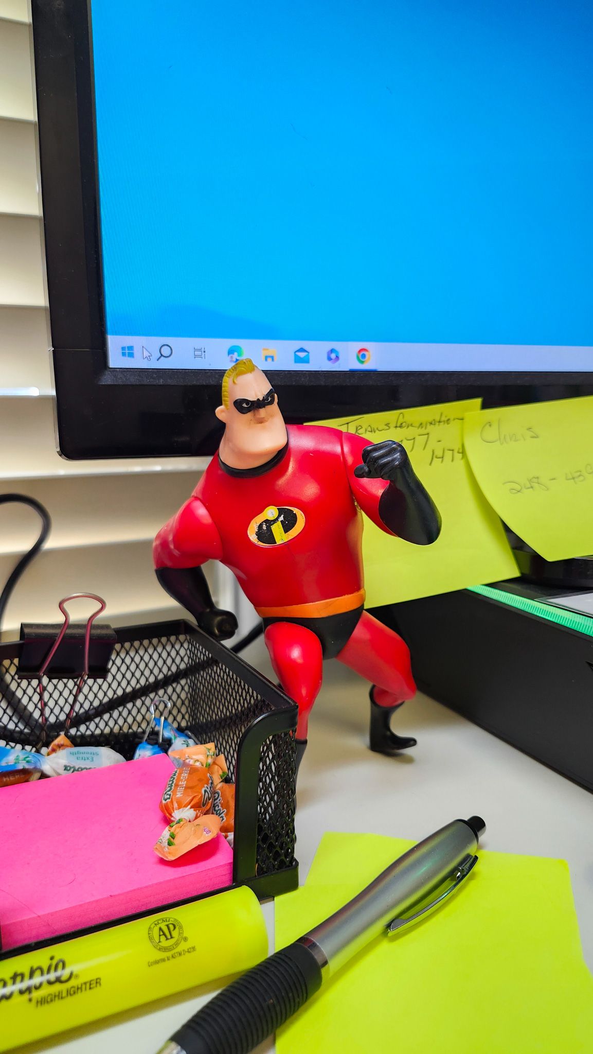 A toy of mr. incredible is sitting on a desk next to a pen and sticky notes.