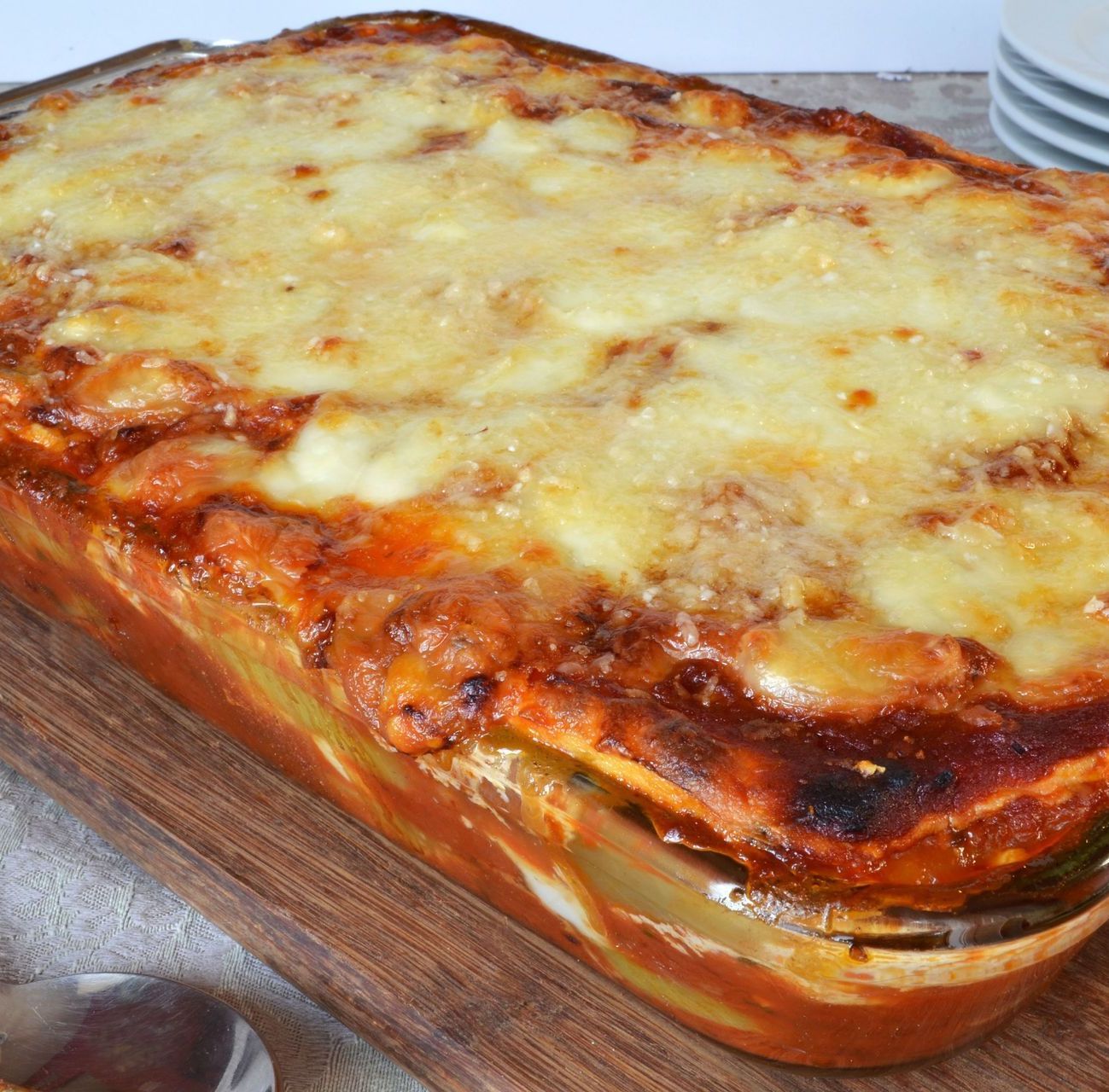 A lasagna is sitting on a wooden cutting board next to a spoon.