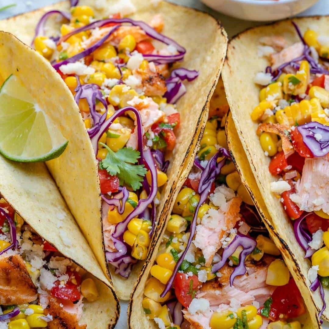 A close up of three tacos with corn , cabbage , and tomatoes on a table.