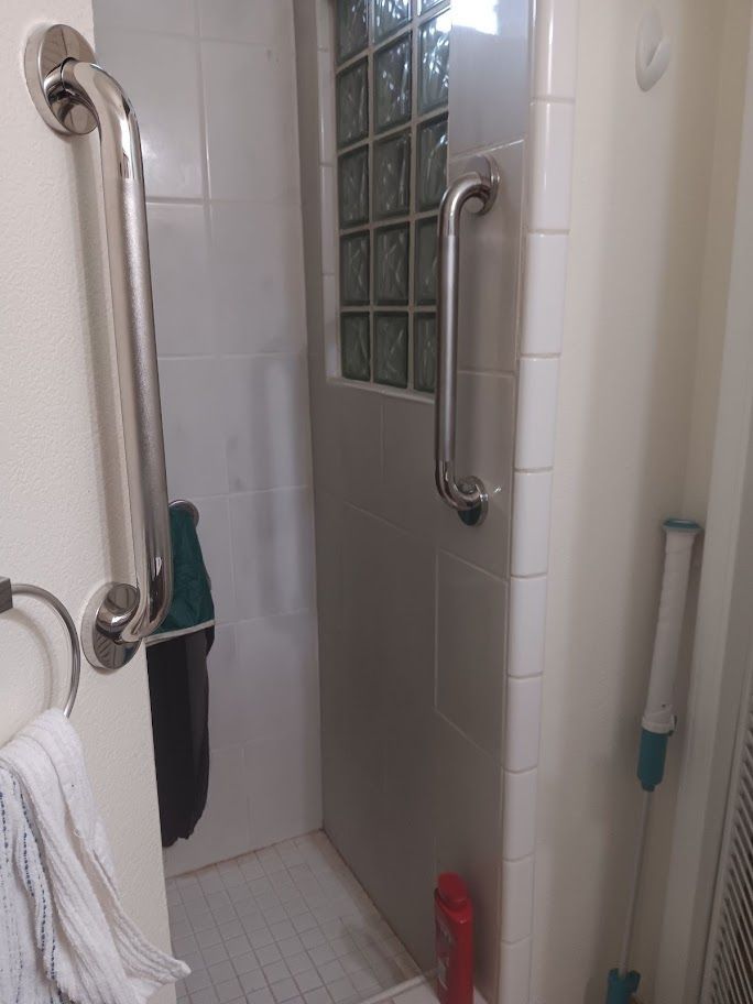 Shower with grab bars, towel ring, and cleaning tools. Light tiles, glass block window, and red bottle visible.