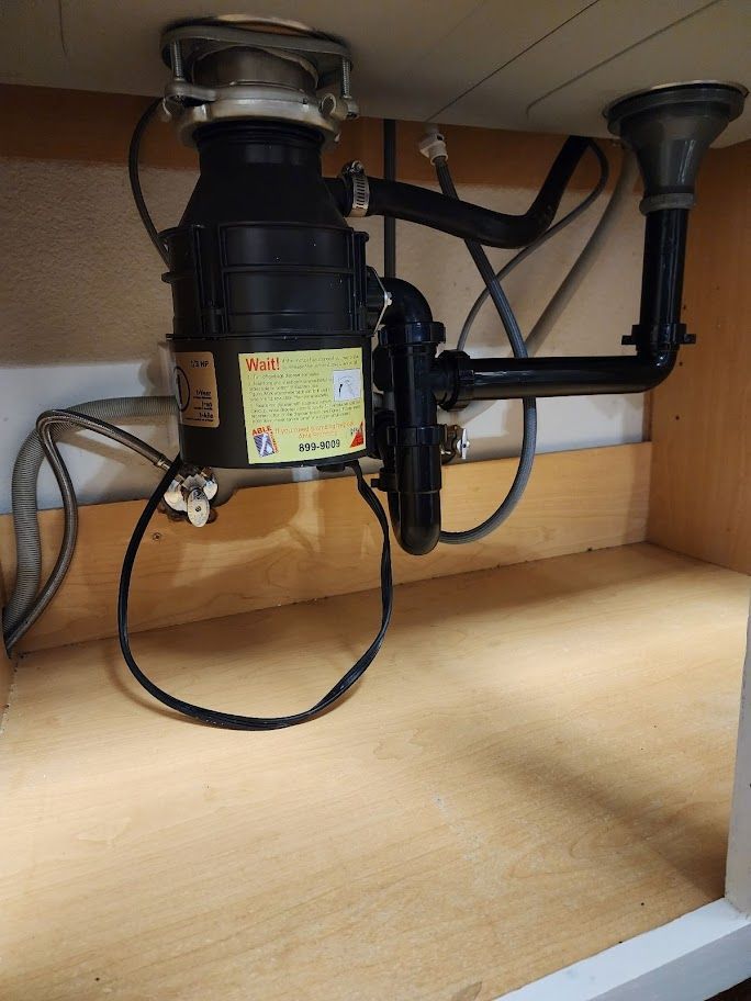Under-sink view of a garbage disposal, plumbing pipes, and sink drain against a wooden cabinet backdrop.