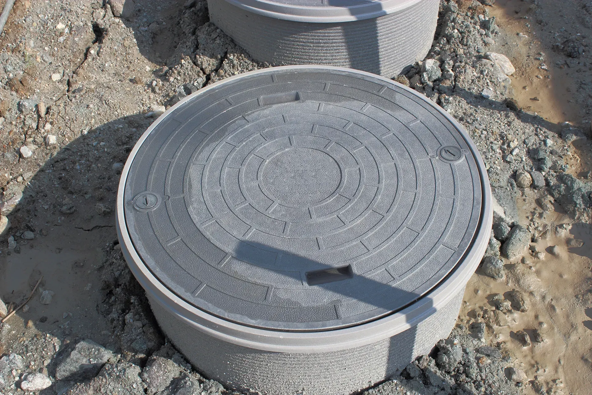 Circular gray concrete utility cover with textured design, set in gravel ground.