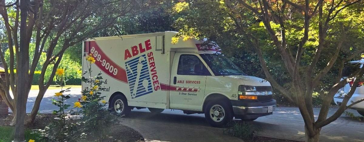 A white plumbing truck is parked in a driveway surrounded by trees.