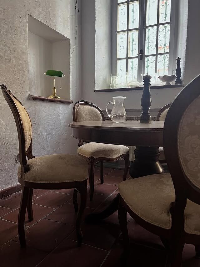 Sala da pranzo con tavolo rotondo, sedie decorate, piccola lampada su nicchia a parete, finestra.