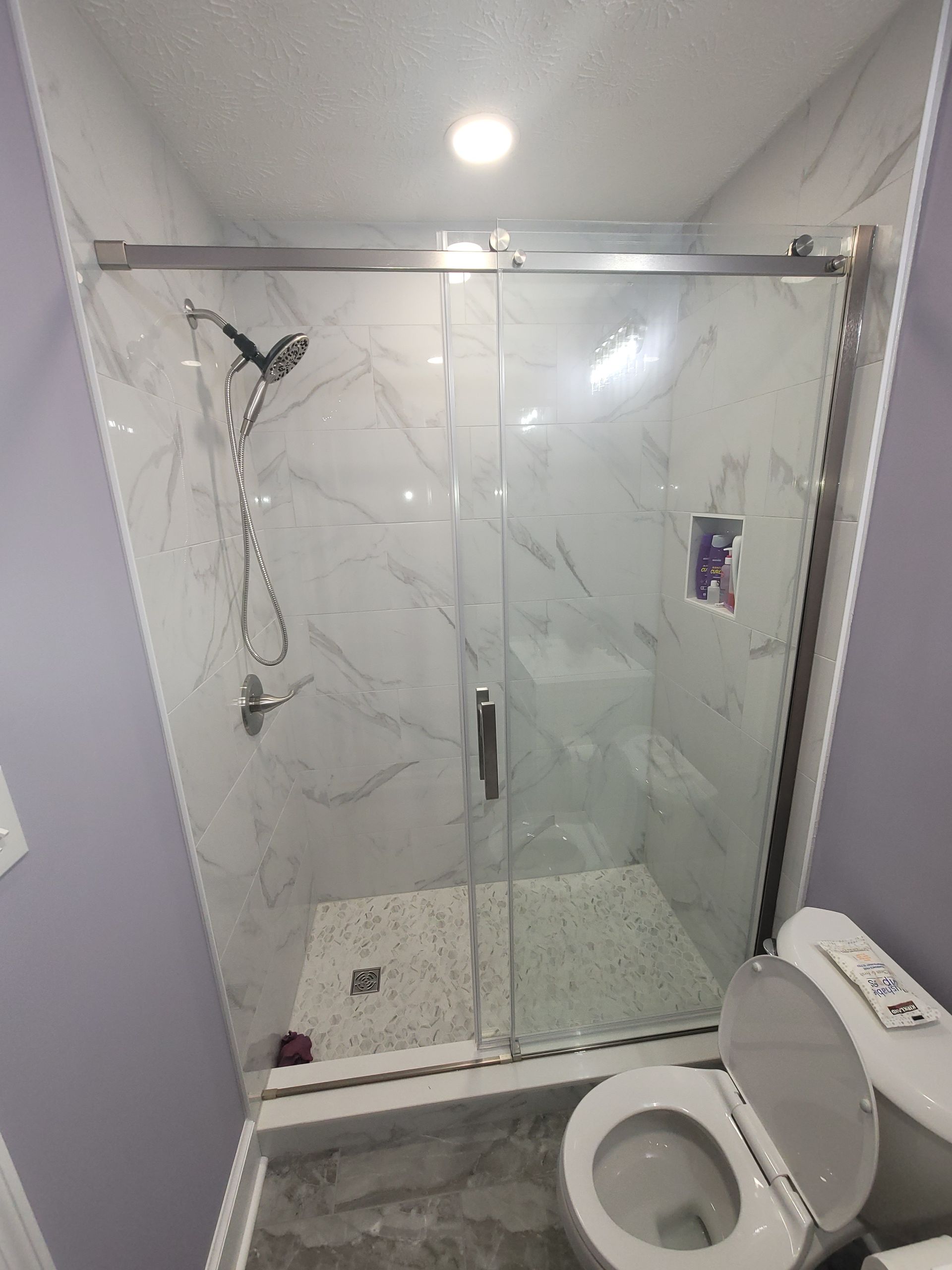 A modern shower with marble-look tile walls, a sliding glass door, and a pebble floor, located next to a white toilet.