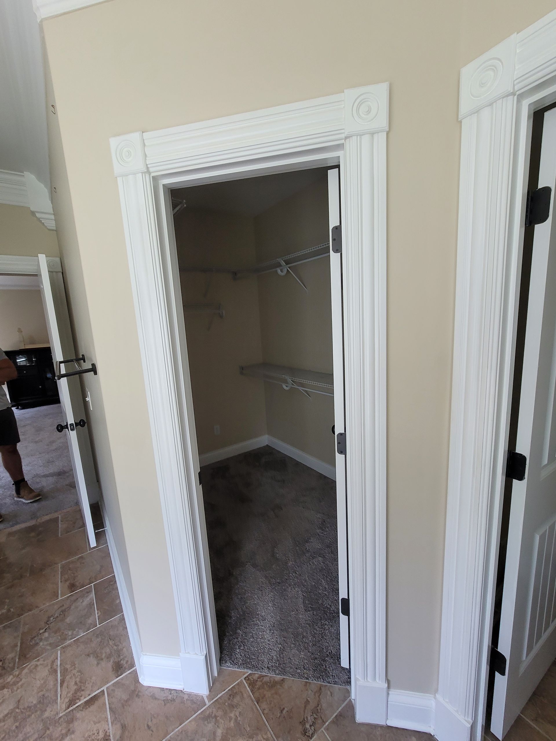 A small walk-in closet with carpet and shelves, framed by decorative white molding, viewed from a tiled hallway.