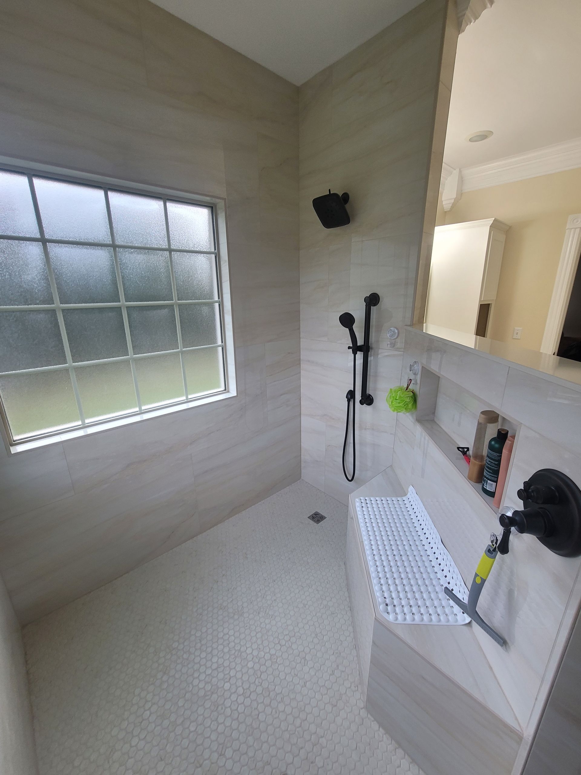 A modern walk-in shower with light tile walls, a pebbled floor, a corner bench, a handheld showerhead, and a window.