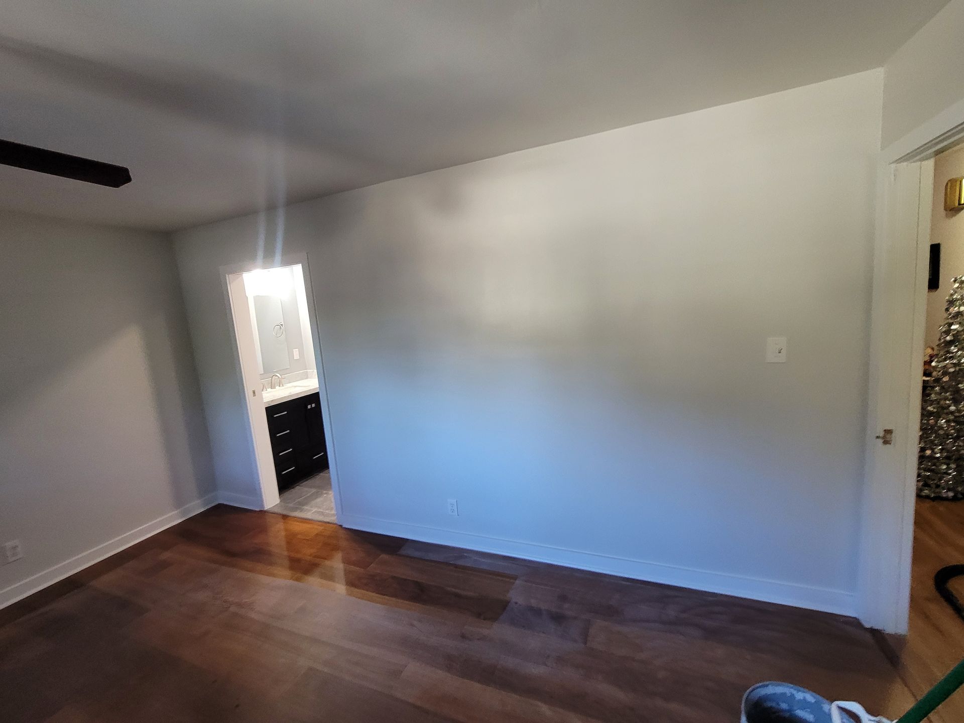 An interior room features a large light-gray wall, dark wood floors, a doorway leading to a bathroom, and white trim.