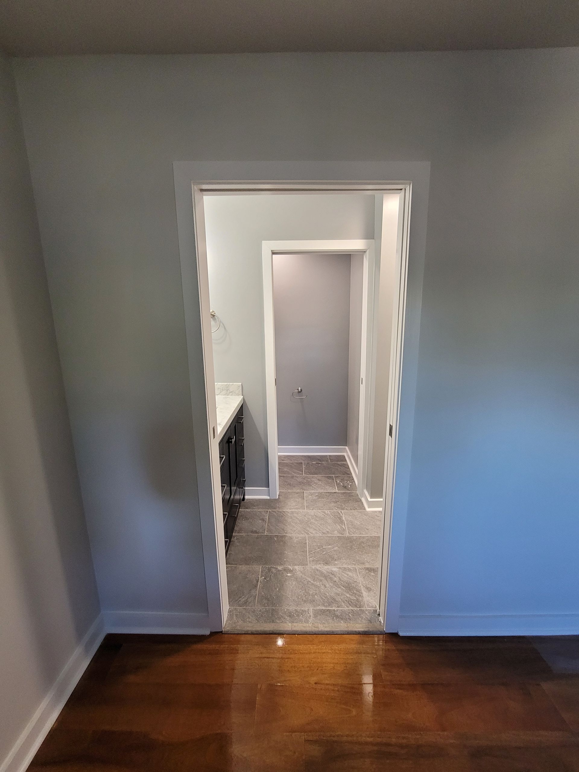 A doorway with white, scalloped trim connects a hardwood-floored room to a bathroom featuring gray walls and stone tiles.