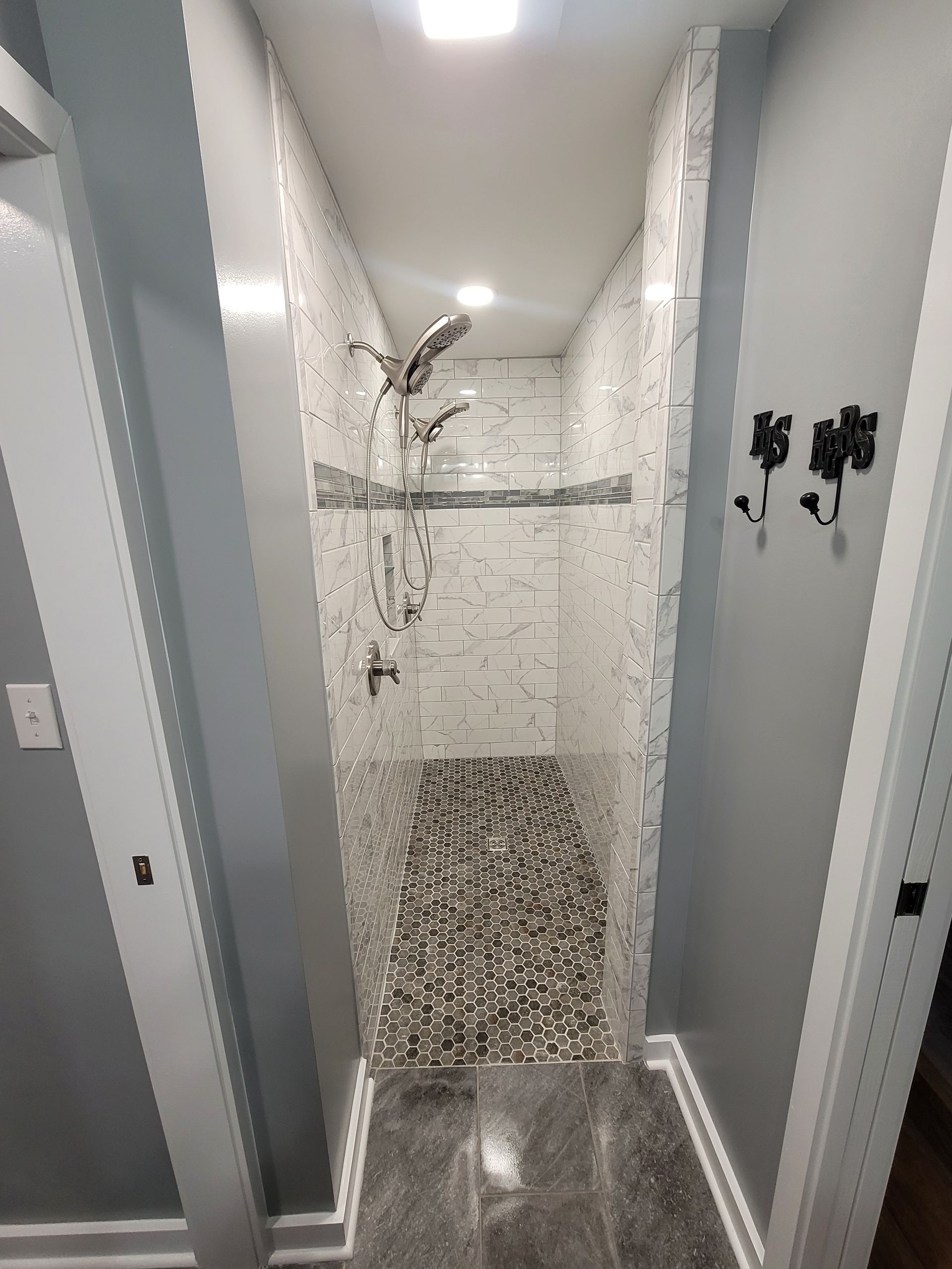 A narrow shower stall with white stone-look tiled walls, a pebble floor, a chrome showerhead, and a glass shelf.
