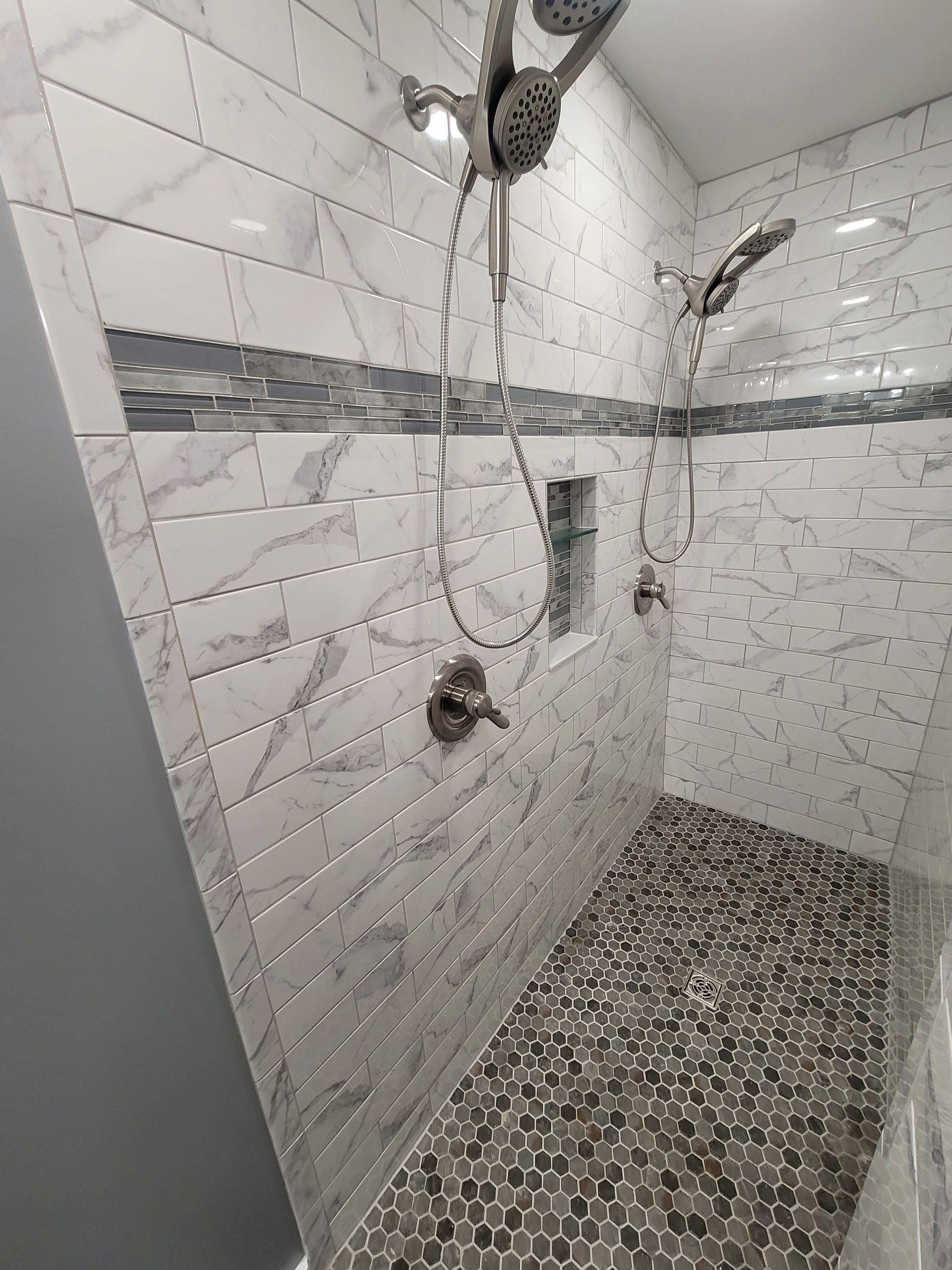 A walk-in shower with white marble-look tiles, a dark pebble floor, a built-in shelf, and brushed nickel shower fixtures.