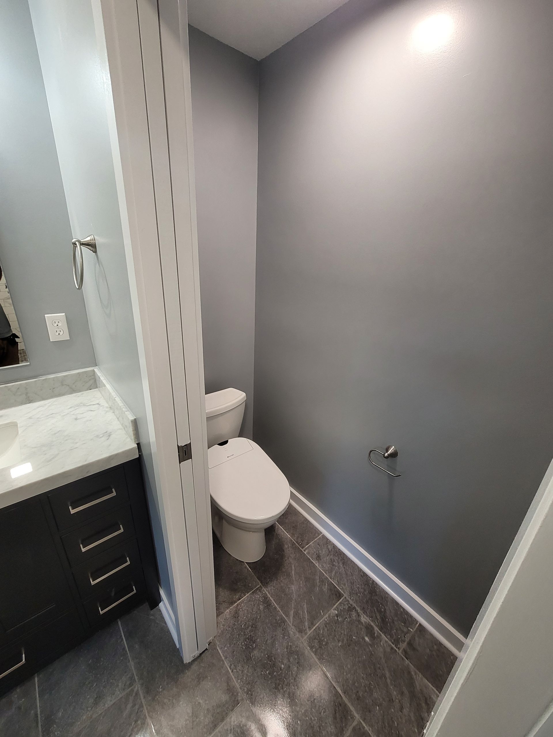 A bathroom with a gray-walled toilet nook, a vanity with a white countertop, and dark gray tiled floors.