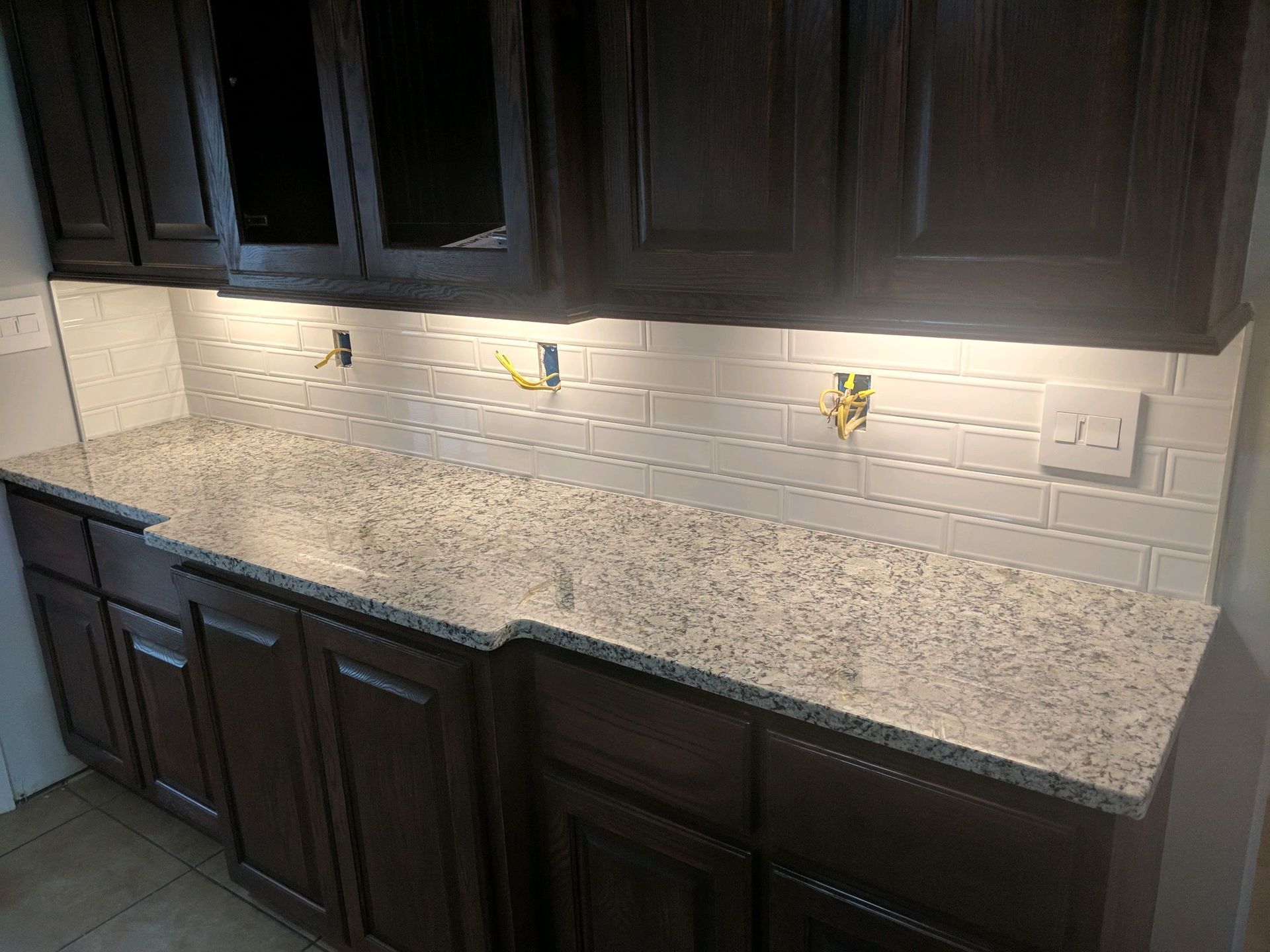 A kitchen counter with speckled granite, dark wood cabinets, and an unfinished white subway tile backsplash.