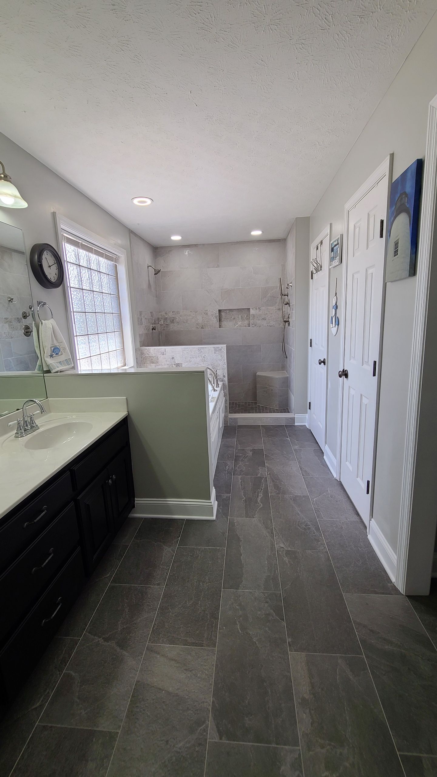 A modern bathroom with dark cabinets, a white countertop, a partial green wall, grey tiled floors, and a walk-in shower.