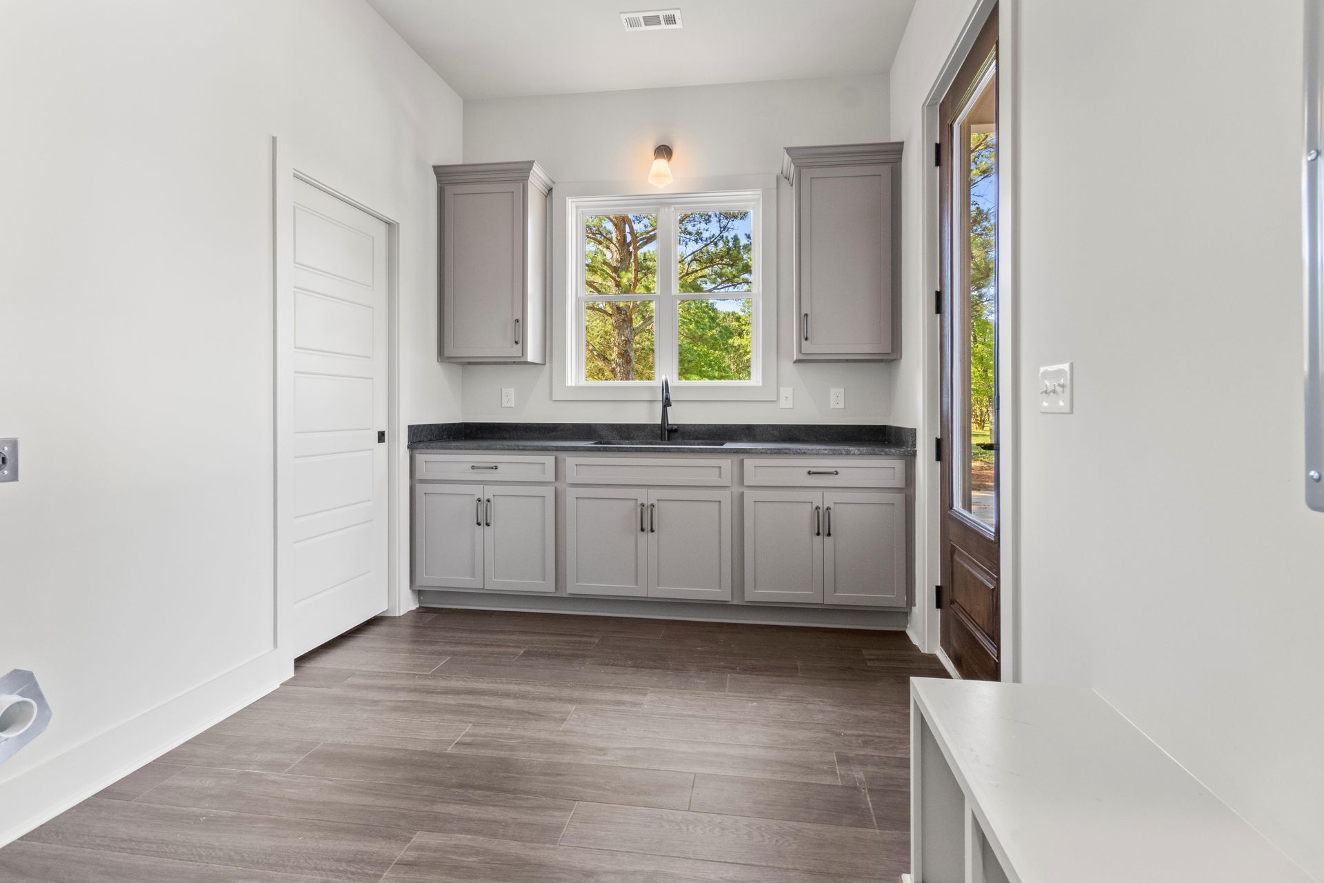 A bright laundry room with gray cabinets, a black countertop, a window, brown wood-look floors, and a white door.