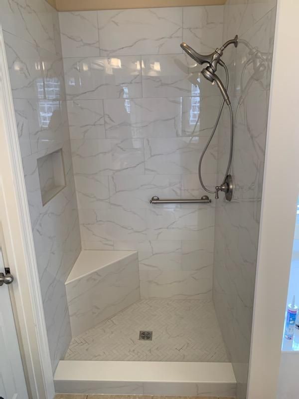 A walk-in shower with white marbled tile, a built-in corner seat, a hand-held shower head, and a horizontal grab bar.