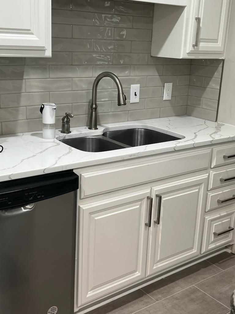 A kitchen sink area with white shaker cabinets, a marble-patterned countertop, grey tile backsplash, and stainless faucet.