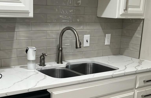 A kitchen sink with a high-neck faucet, mounted on white marble-style countertops against a gray subway-tiled backsplash.