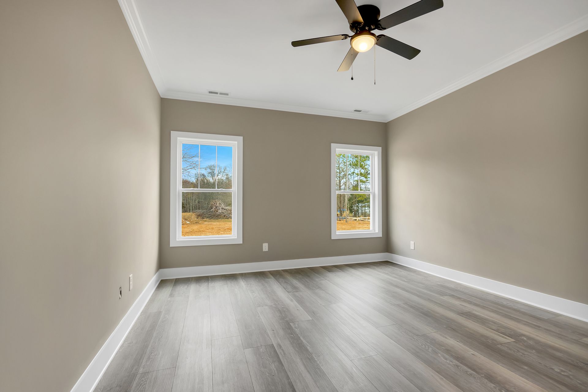An empty room with light beige walls, gray wood-look flooring, two windows, and a ceiling fan.