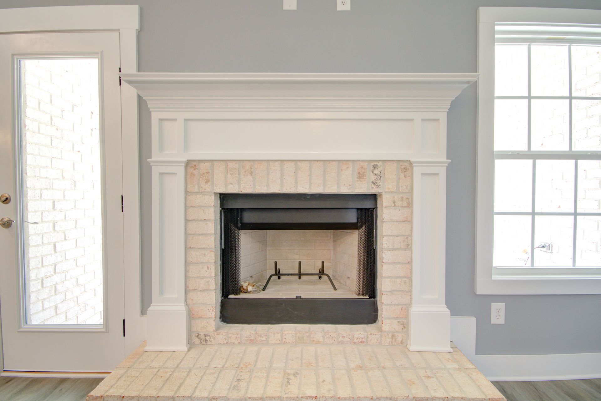 A white fireplace mantel with a light-colored brick surround and hearth, set against a soft gray wall in a room.