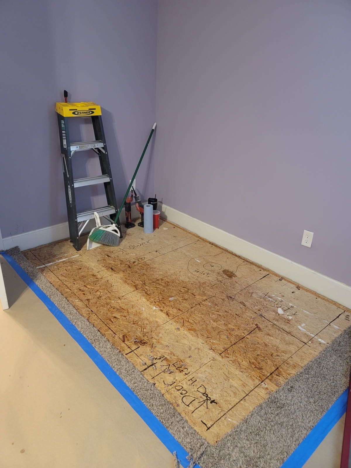 A room corner with a step-up floor platform featuring exposed plywood underlayment, a ladder, and painter's tape.