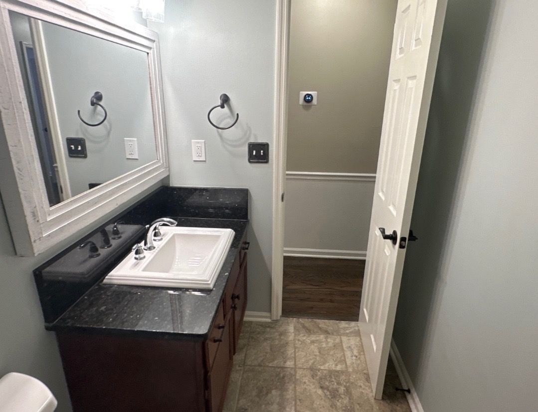 A bathroom vanity with a black countertop and white sink, white-framed mirror, and an open door to a room with wood floors.