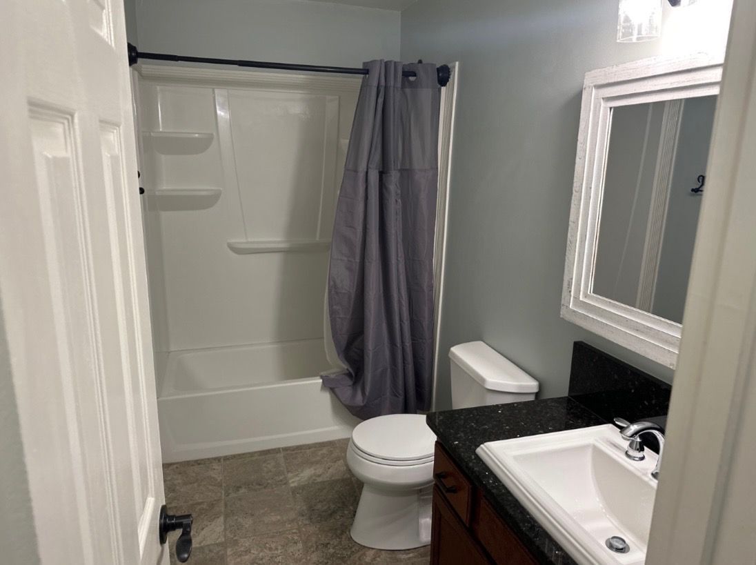 A bathroom with a white tub/shower, grey curtain, white toilet, dark vanity with a white sink, and a framed mirror.