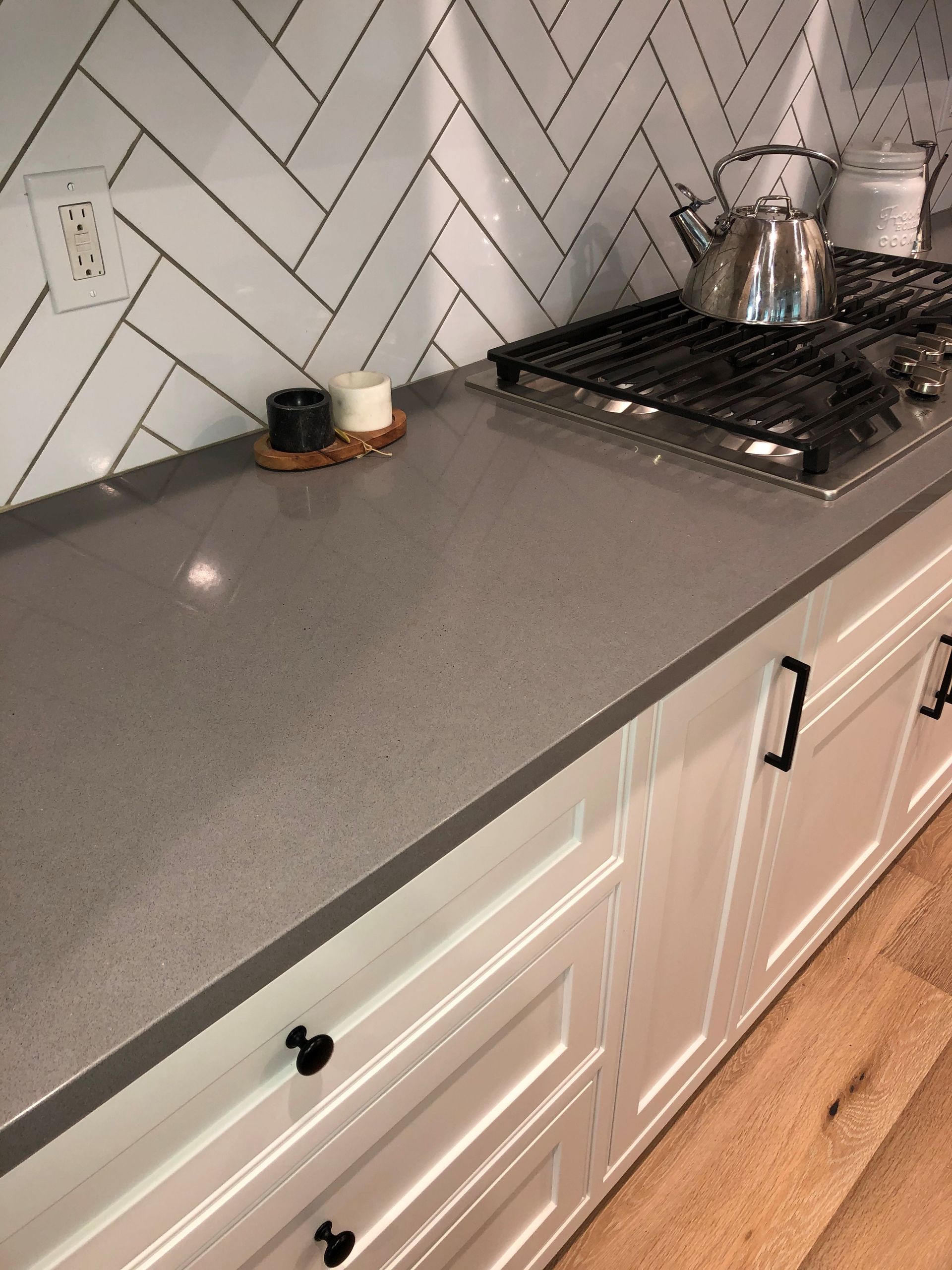 A kitchen counter with a gray quartz top, white herringbone tile backsplash, and white cabinets with black hardware.