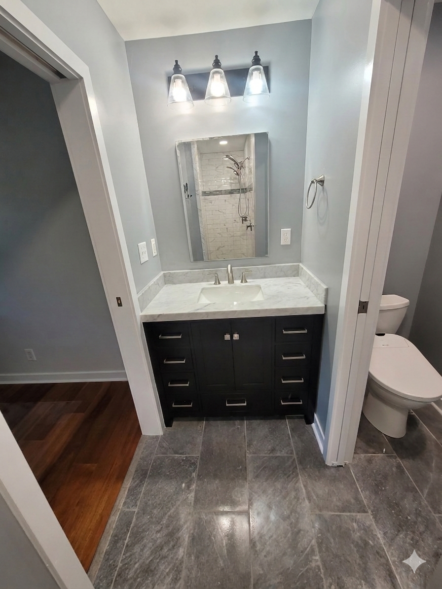 A bathroom with a dark wood vanity, white countertop, rectangular mirror, three-light fixture, and grey tiled floors.