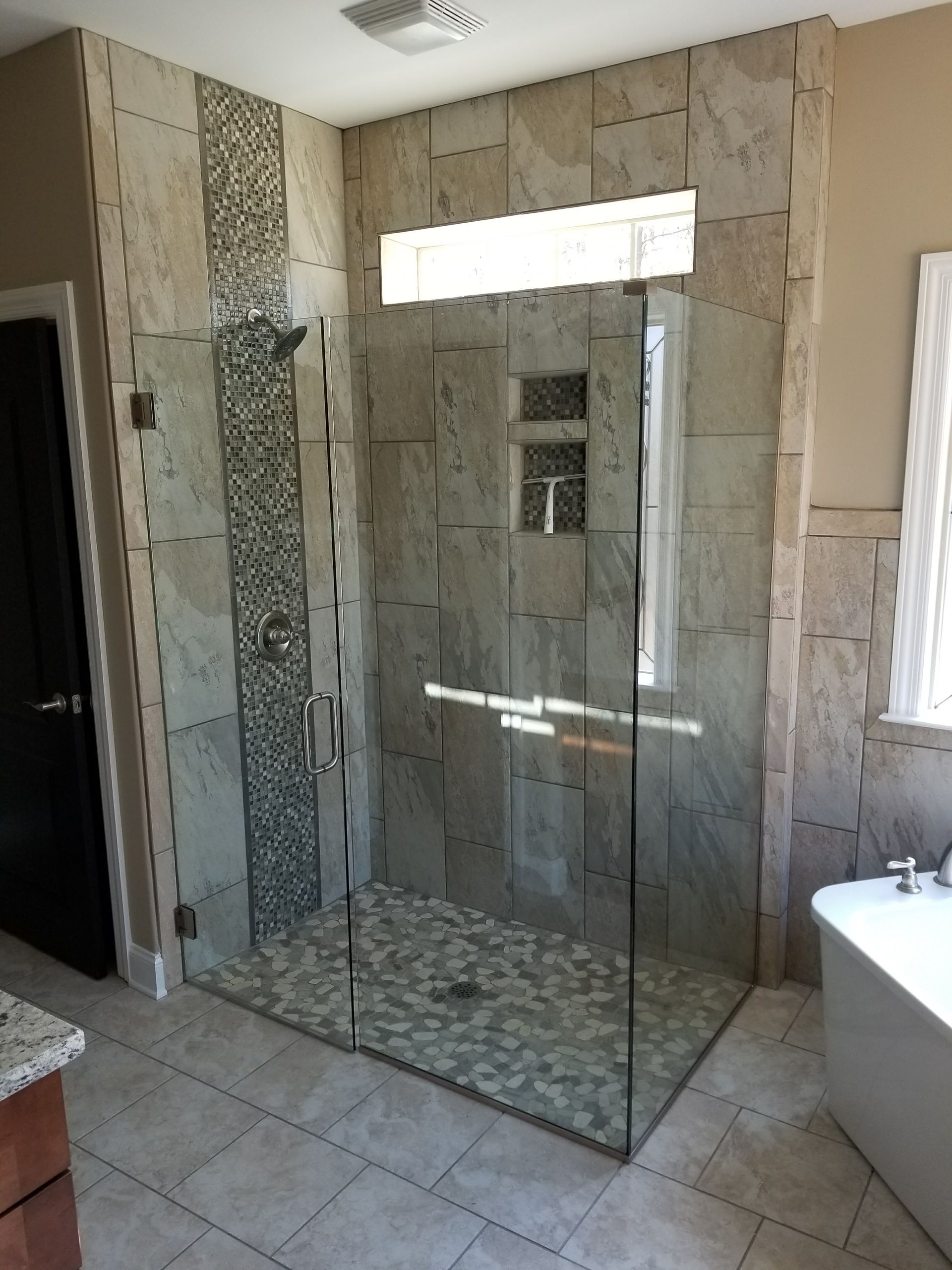 A walk-in shower with a glass door, natural stone tile walls, a mosaic vertical accent strip, and pebble tile flooring.