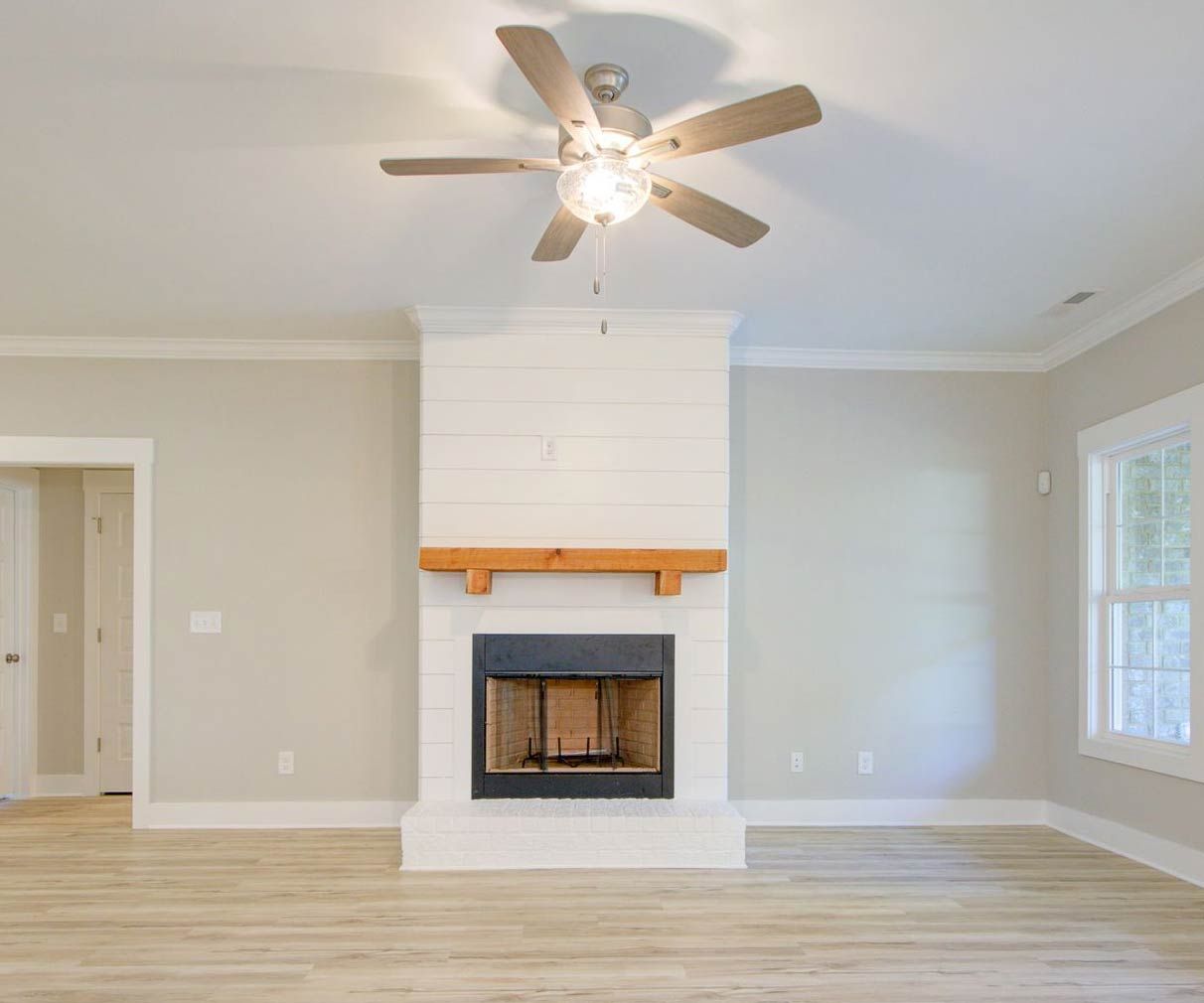A bright, minimalist room features light wood floors, a white shiplap fireplace with a wood mantel, and a ceiling fan.