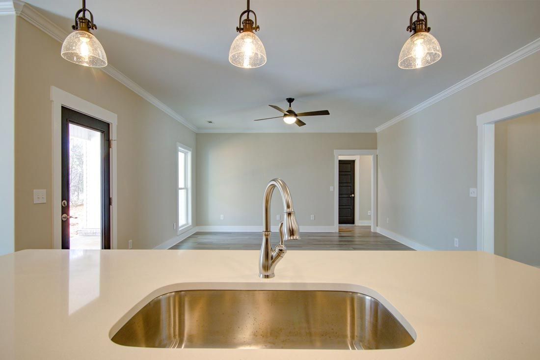 A modern kitchen island with a metal sink and faucet, looking into an open-plan living area with pendant lights.