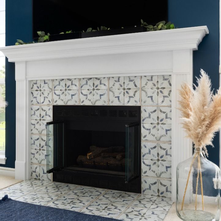 A fireplace with white mantel and blue-and-white patterned tile surround, accented by a vase of dried pampas grass.