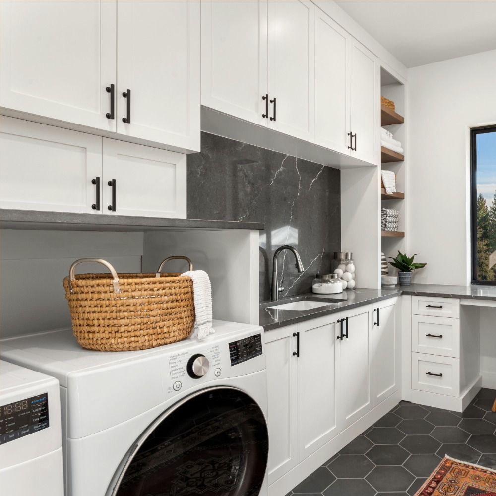 A modern laundry room with white cabinets, a black marble backsplash, dark hexagon floor tiles, and a wicker basket.